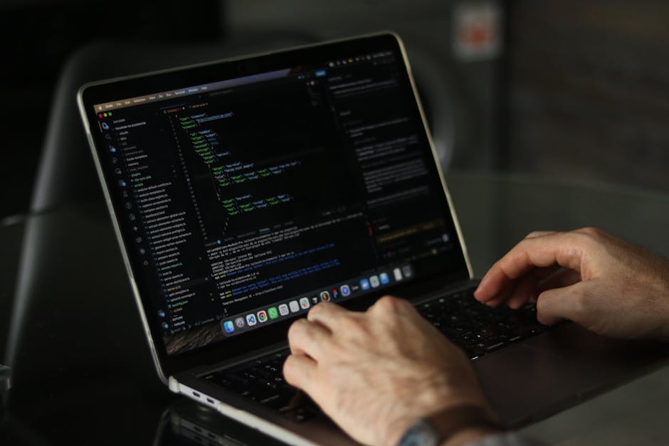 A person with light skin and visible arm tattoos is working on a MacBook laptop placed on a dark surface, possibly a table. The screen displays a code editor with lines of colorful code, including gre